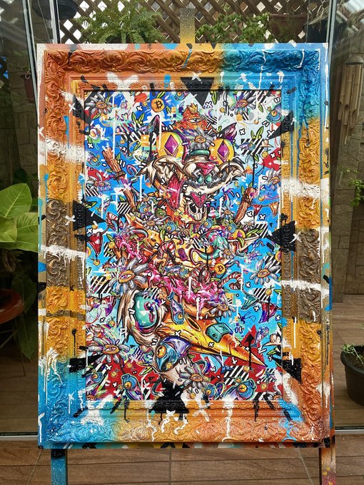A vibrant, abstract painting on canvas featuring a colorful array of symbols, including a bee, crown, skull, and geometric patterns in bright blues, oranges, and reds. The artwork is displayed outdoors against a backdrop of green foliage.