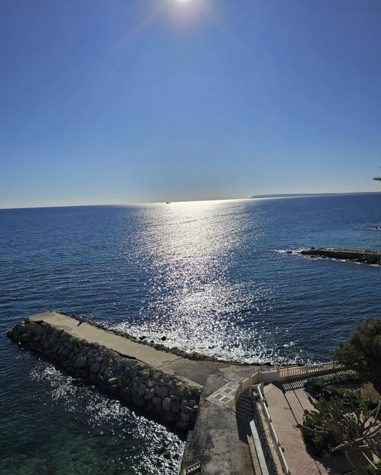 The image shows a beautiful view of the ocean with a pier extending into the water. The sun is shining brightly, creating a stunning reflection on the surface of the water. There are also some people visible near the pier, enjoying their time by the water.