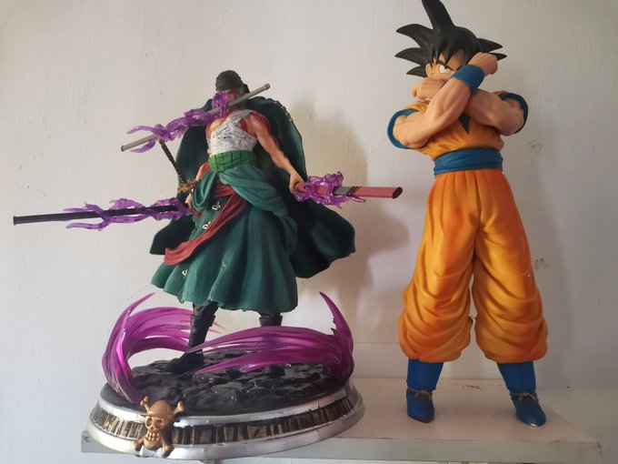 Two anime figurines displayed on a shelf: a green-robed sword character with purple energy effects on the left, and Goku in his signature orange gi on the right.