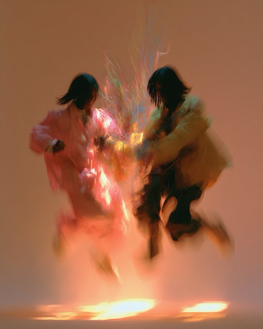 An artistic, blurred image of two figures in flowing garments, one in pink and one in yellow, appearing to dance or leap against a warm orange background.