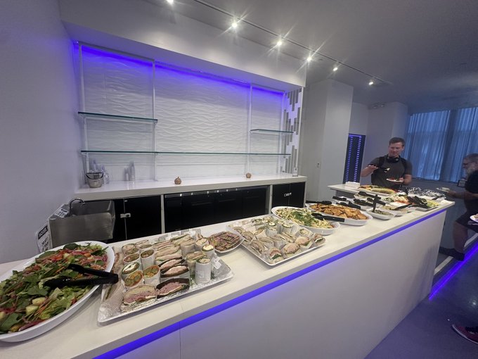A buffet spread featuring sandwiches, salads, and pastries on a white counter with purple LED lighting, where a person in black is serving themselves food.