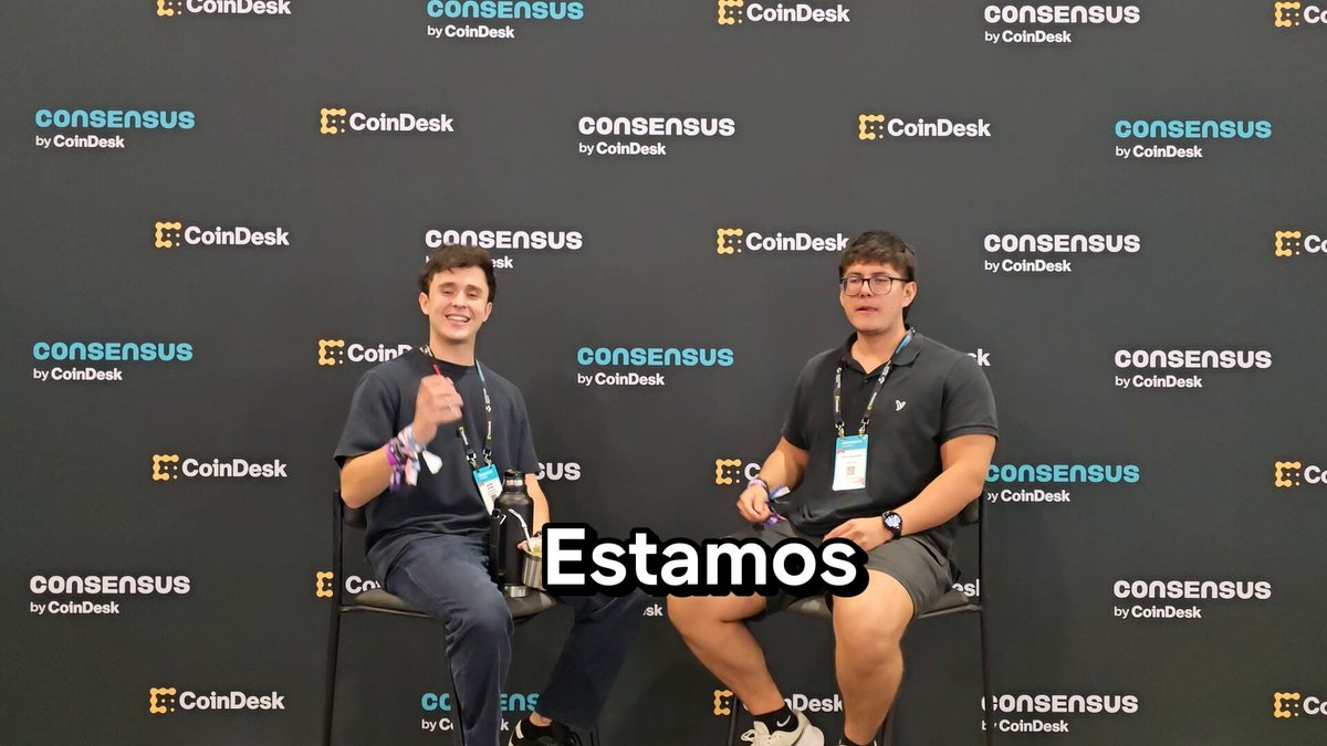 The image shows two men sitting next to each other with their arms crossed, both wearing black shirts and holding controllers. They are positioned against a backdrop of a large screen displaying text related to tokenization and agents. The text reads "From HederaCon to tokenization and agents, builders sharing what they're learning and building in real time." This suggests that the men might be discussing or participating in an event or conference related to cryptocurrency development and tokenization.