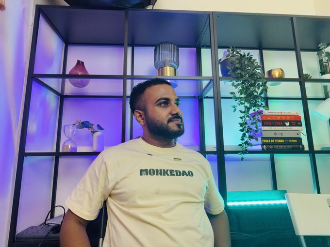 A man wearing a white 'MONKEDAO' t-shirt sits in front of a black shelving unit with purple and teal lighting, decorative plants, and books in the background.