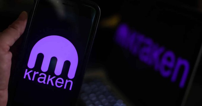 A smartphone displaying the purple Kraken logo on its screen, held by a hand in front of a laptop.