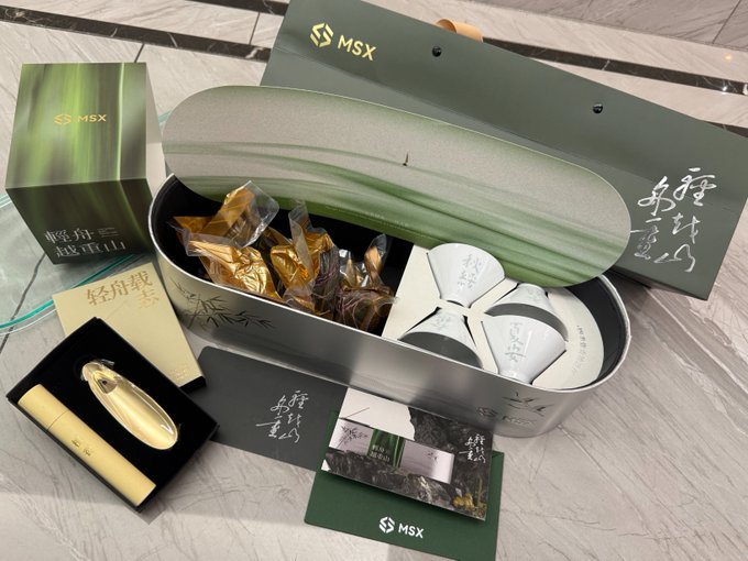 The image features a silver box with a green lid, containing various items such as tea cups, a hair dryer, and other accessories. The box also includes some papers that are not clear about their contents or brand names. The overall scene suggests that these items might be part of a gift set or a promotional offer for the MSX brand.
