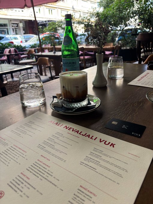 A bottle of Pellegrino sparkling water and an iced coffee sit on a wooden café table alongside a menu titled "NEVALJALI VUK" (БАР / BAR RESTORAN), capturing a relaxed morning breakfast scene in Belgrade that matches the post's mention of a great coffee break with local friends before heading into day two of the Solana Summit.