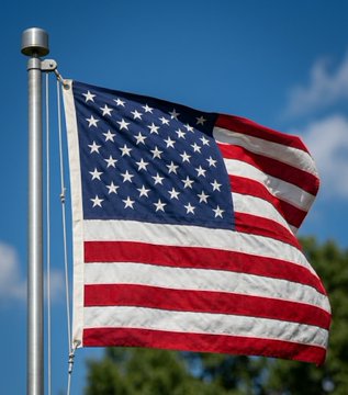 The image features a large American flag flying high against a blue sky with white clouds. The flag has red, white, and blue stripes that represent the colors of the United States. The flag is attached to a metal pole at an angle, indicating its height above the ground.