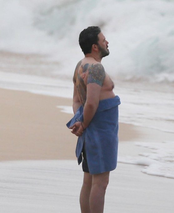 Ben Affleck stands on a beach with a blue towel wrapped around his waist, displaying arm tattoos as he looks out toward the ocean waves.