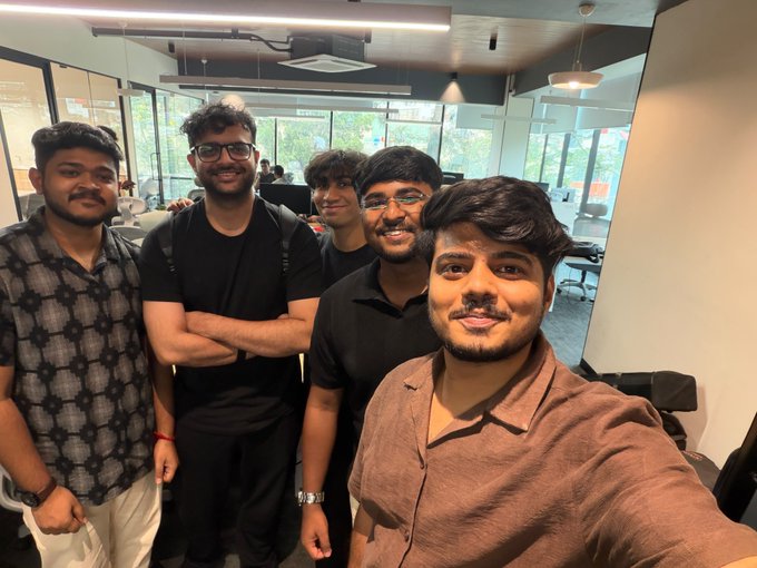 A group of five young men smiling for a selfie in a modern office space, with one man in a brown shirt taking the photo in the foreground.