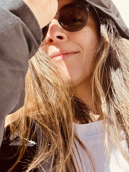 A close-up selfie of a smiling young woman with long blonde hair, wearing sunglasses and a dark hooded jacket, photographed in bright sunlight.