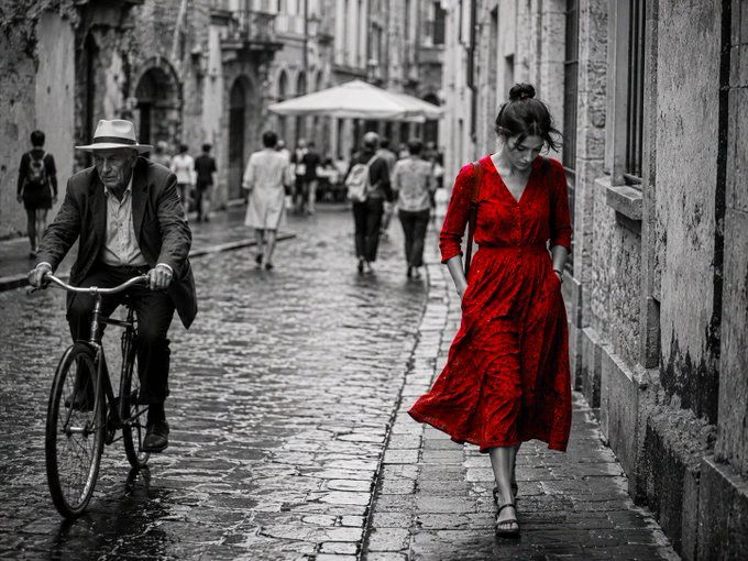 A black-and-white street photograph featuring a woman in a red dress walking down a cobblestone alley, while a man in a suit rides a bicycle on the left.