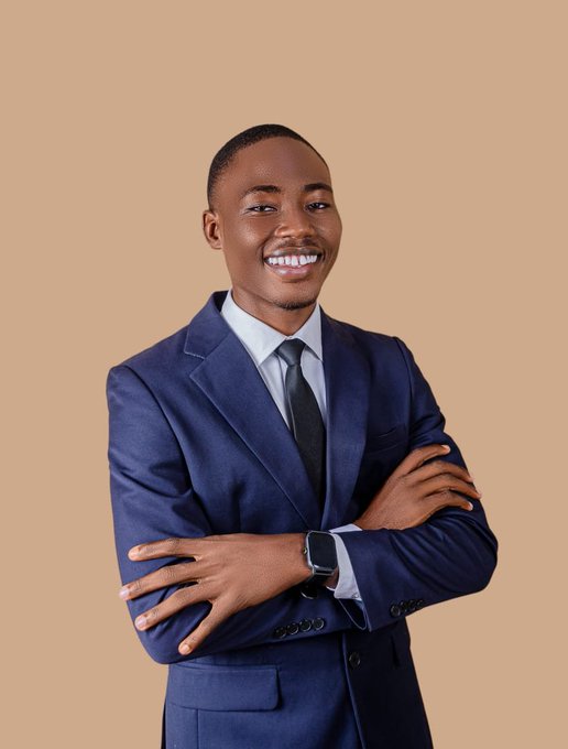 A smiling young man wearing a navy blue suit, white shirt, and black tie stands with crossed arms against a beige background.