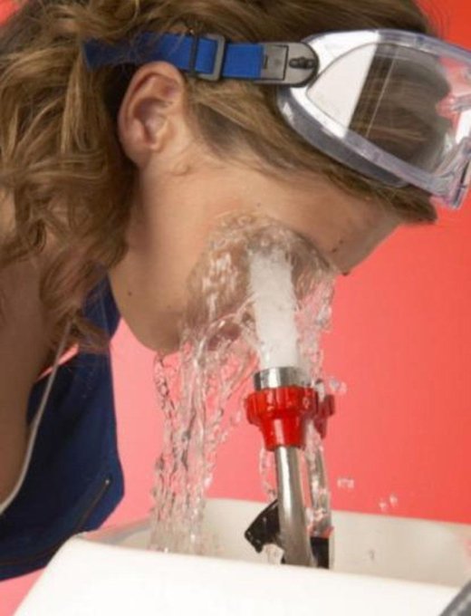 A child wearing protective safety goggles washes their face at a water fountain, with water splashing around their face against a red background.