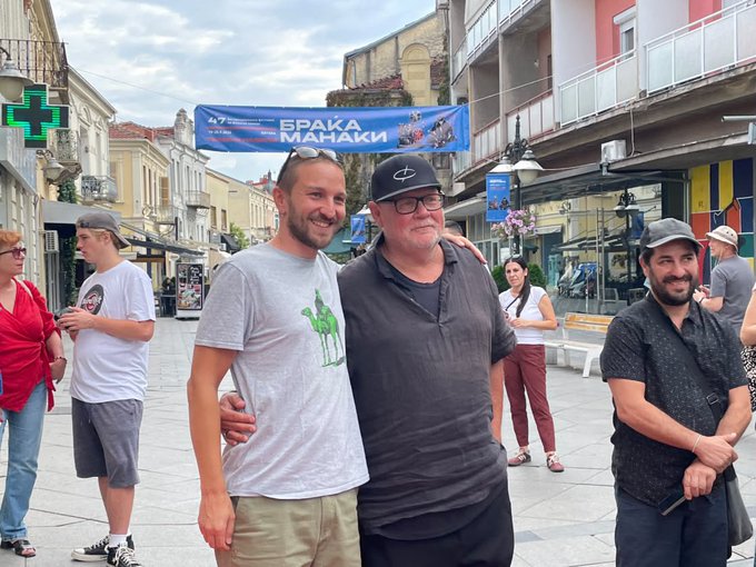 A street photo of three smiling men posing together on a pedestrian street with a banner reading 'БРАКА МАНАКИ' and a green cross pharmacy sign visible in the background.