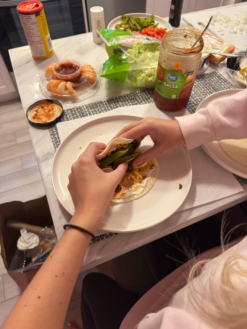 A person assembles a taco on a plate surrounded by taco ingredients, with a Kroger brand Salsa jar prominently visible. The scene captures a homemade Taco Tuesday celebration, connecting the celebratory post with the hands-on, grateful cooking effort shown.