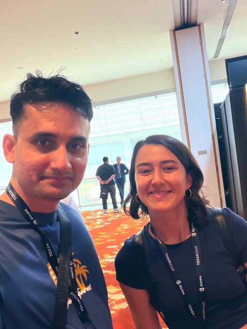 A selfie of a man and a woman smiling at the camera at a TOKEN2049 conference, both wearing lanyards and dark shirts.