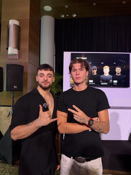 Two stylishly dressed young men pose confidently in front of a television screen displaying what appears to be their interview, with one wearing a black button-up shirt and the other sporting a black tee and white pants, both making hand gestures as they greet followers with a casual "GM" (Good Morning).