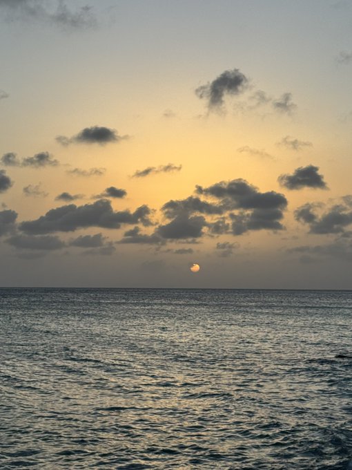 A calm ocean stretches to the horizon where the sun rises (or sets) through scattered clouds, casting a soft golden glow over the rippling water—offering a serene, hopeful "Good Morning" to greet the day.
