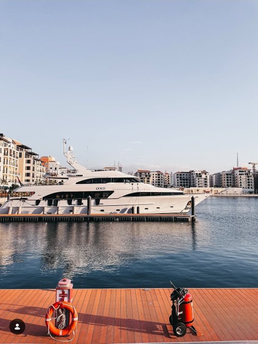 A large white luxury yacht docked at a marina in a city with modern apartment buildings in the background, viewed from a wooden deck where a life ring and fire extinguisher are visible in the foreground.