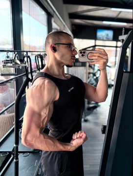 A muscular man in a black tank top and glasses takes a mirror selfie with his phone inside a gym.
