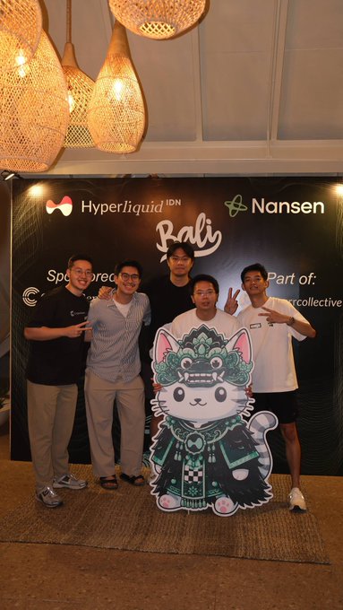 Five young men pose in front of a banner for 'Hyperliquid | IDN Nansen Bali Sponsors' with a mascot, under decorative woven light fixtures.