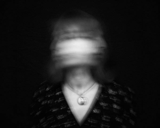 A blurred black and white portrait of a person wearing a patterned dark top and a circular pendant necklace, with motion blur obscuring their facial features.