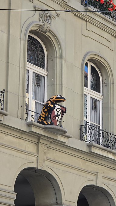 A whimsical frog sculpture perches on the wrought-iron balcony of an ornate building in Bern, Switzerland, blending playful street art with the city's elegant Belle Époque architecture. The large toad figurine holds a shield, standing out vividly against the pale stone façade and arched windows.