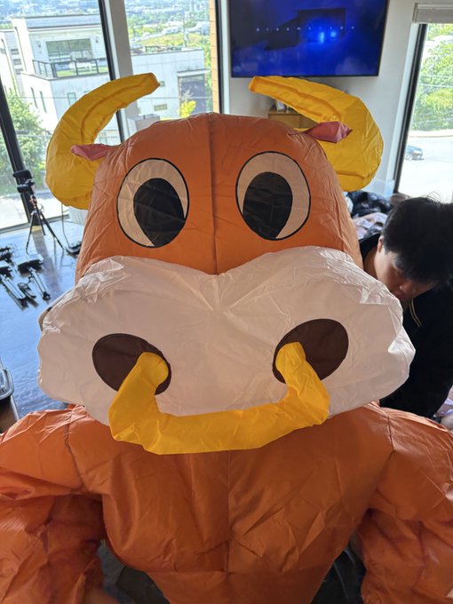 An inflatable orange bull mascot with large cartoonish eyes and yellow horns is displayed indoors, with a person visible in the background adjusting the costume. The image relates to "$ANSEM takeover" by embodying the bullish energy suggested by the stock reference, with the inflatable bull serving as a visual symbol of a strong market takeover.