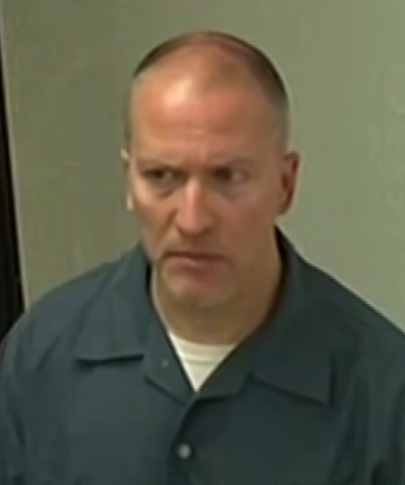 A man with a shaved head wearing a dark teal prison-style shirt over a white undershirt stands against a plain gray wall.