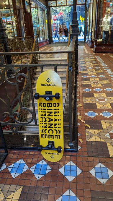 A bright yellow skateboard branded with "BINANCE" branding leans against an ornate wrought-iron railing inside a Victorian-style arcade with tiled floors. The juxtaposition of the modern crypto-branded board in the historic shopping arcade visually represents Binance's effort to bring its digital ecosystem into real-world, offline spaces.
