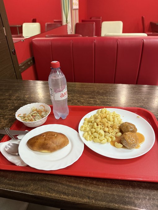 A red tray on a restaurant table holds a meal of pasta with meatballs, an empanada, a side dish, and a plastic water bottle, set against red booth seating.