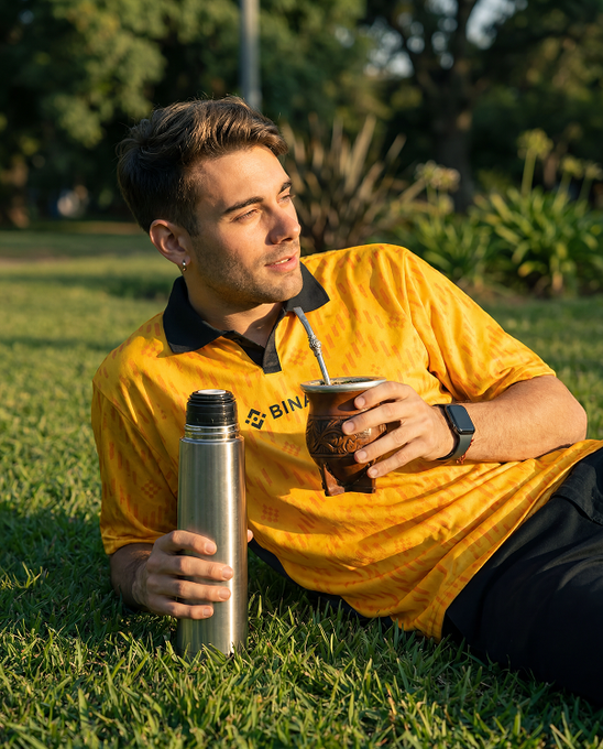 A young man lounges on grass in a yellow jersey partially showing "BINA" on the chest, holding a stainless steel thermos and a traditional mate cup with a bombilla, fitting the post's "ready for kickoff" vibe of South American game-day leisure.