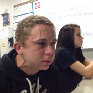 A young man in a black hoodie looks solemnly into the lens with a defeated, dead-inside expression in a classroom setting. Behind him, a whiteboard displays partially visible text including "No subscribers" and "Active Subs," humorously reflecting the post's lament about the agonizing wait between scans of low-subscriber Minecraft YouTubers.