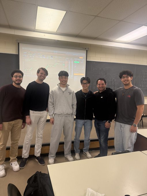 A group of six young men standing together smiling in a classroom with a projected financial chart displayed on the screen behind them.