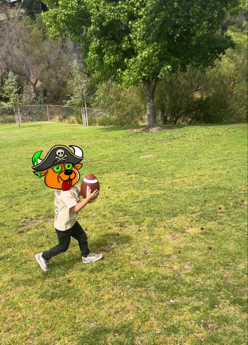 A young child running across a grassy field holding an American football, with their face obscured by a cartoon pirate dog filter.