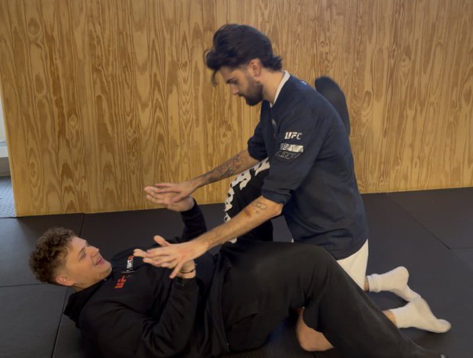 Two men in UFC apparel practice grappling on black mats, with the standing instructor (wearing a shirt reading "UFC") guiding the supine trainee through a technique, humorously symbolizing a "submission hold" on the market as Solana memecoins return.