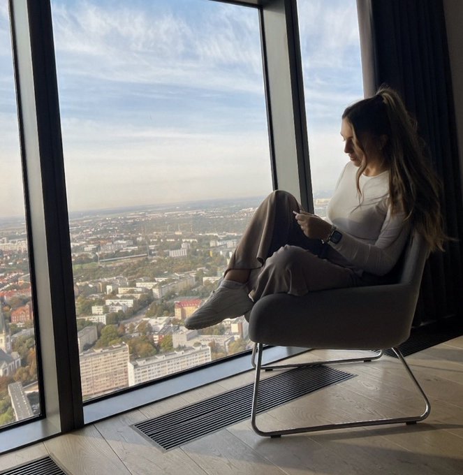 A woman with a ponytail sits in a gray chair by a large floor-to-ceiling window, looking at her phone while overlooking a sprawling cityscape.