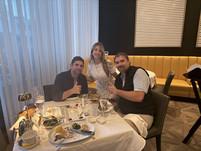 A photograph of three people—a woman in white standing and two men (one giving a thumbs up)—posing at a restaurant table with plates of food, wine glasses, and a candelabra.