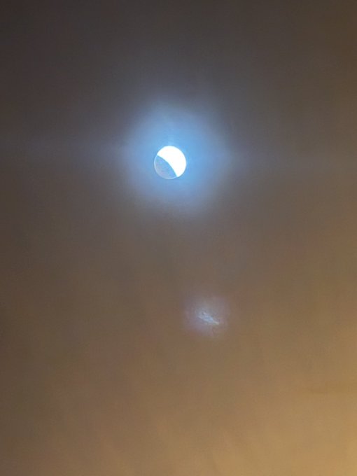 A bright moon shines through clouds at night, with a smaller light source visible below it.