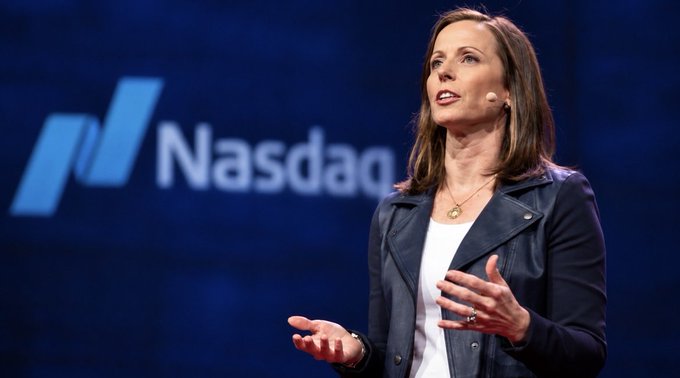 A woman in a dark blazer and white top speaks at a podium, gesturing with her hands, against a blue background displaying the 'Nasdaq' logo.