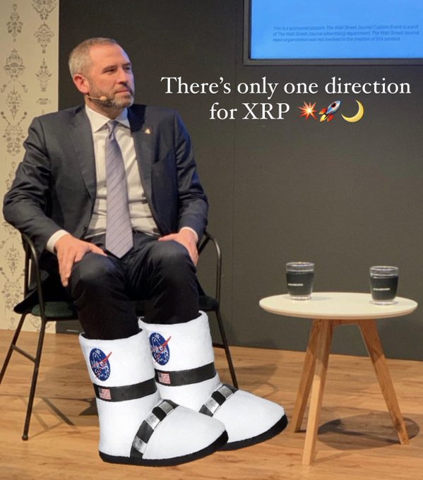 A man in a suit sits on a chair with novelty NASA-themed space boots on his feet, with text overlay reading 'There's only one direction for XRP 🚀🌙' next to a small table with two glasses.