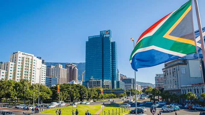 A cityscape photo of Cape Town featuring the South African flag prominently in the foreground, with the FNB building and other downtown structures under a clear blue sky.