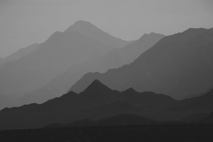 A black-and-white photograph shows layered, hazy mountain silhouettes fading into atmospheric distance, with the nearest dark ridge prominent in the foreground and faint triangular peaks behind.