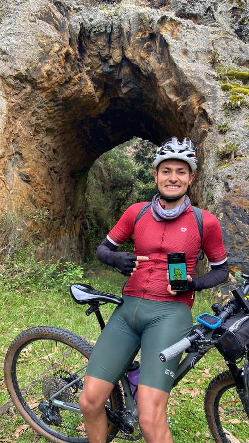 A cyclist in a red jersey and green shorts stands with a mountain bike, holding a phone with a green dinosaur image, in front of a large rock arch.