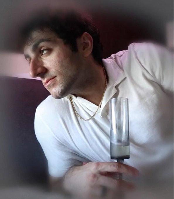 A young man in a white polo shirt and gold chain is seen in profile, holding a tall empty champagne glass while gazing off to the side.