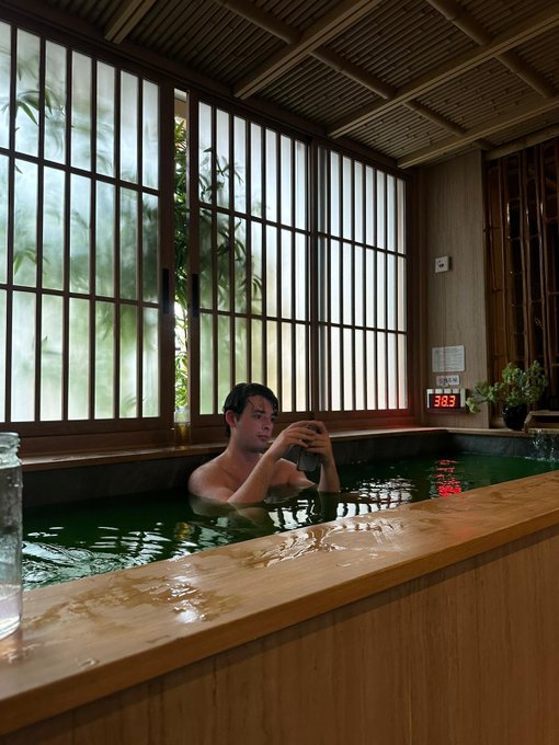 A man relaxes in a wooden Japanese-style hot bath, looking at his phone, with large slatted windows showing bamboo plants in the background.