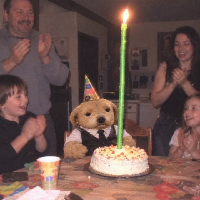 A candid birthday photo showing a teddy bear wearing a party hat seated in front of a cake with an extremely tall green candle, surrounded by clapping family members including children and adults.