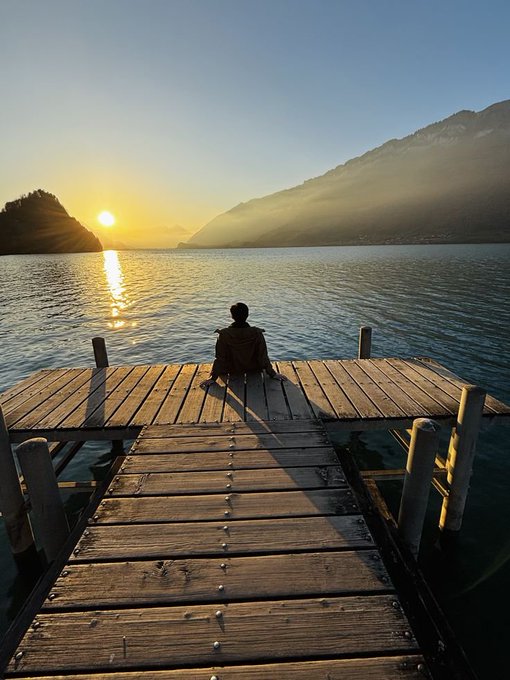 A person sits at the end of a wooden dock on a calm lake, watching a golden sunrise between distant mountains.