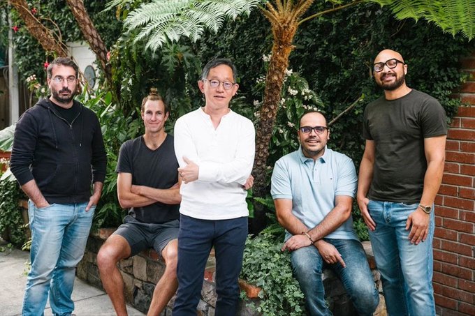 The image shows a group of five men posing together outdoors. They are standing next to each other with their arms crossed, giving off an air of camaraderie or friendship. The background features a brick wall and some plants, adding to the casual atmosphere of the scene. The photo is captioned "building together since @Meta libra, in it for the long game", which suggests that these men have been friends or acquaintances for quite some time and are enjoying each other's company.