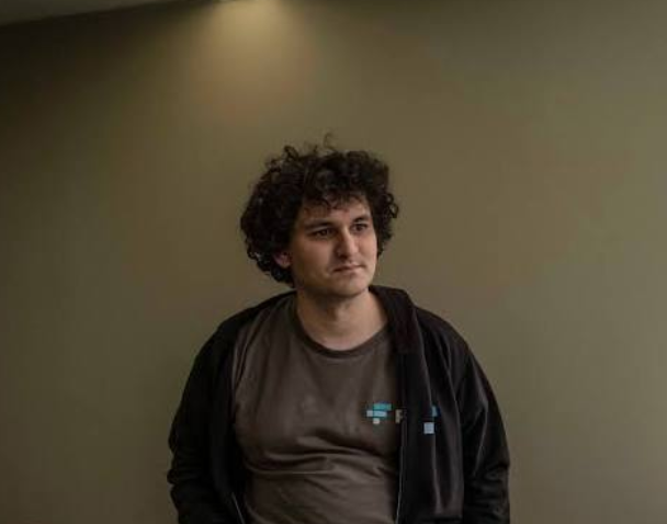 A young man with curly dark hair stands against a plain beige wall under a single light, wearing a dark grey jacket over a brown t-shirt in what appears to be a video still.